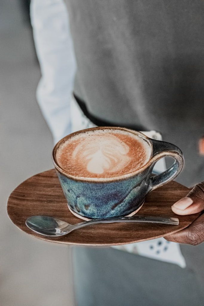 A barista presents a cappuccino with heart-shaped latte art in a blue ceramic cup.